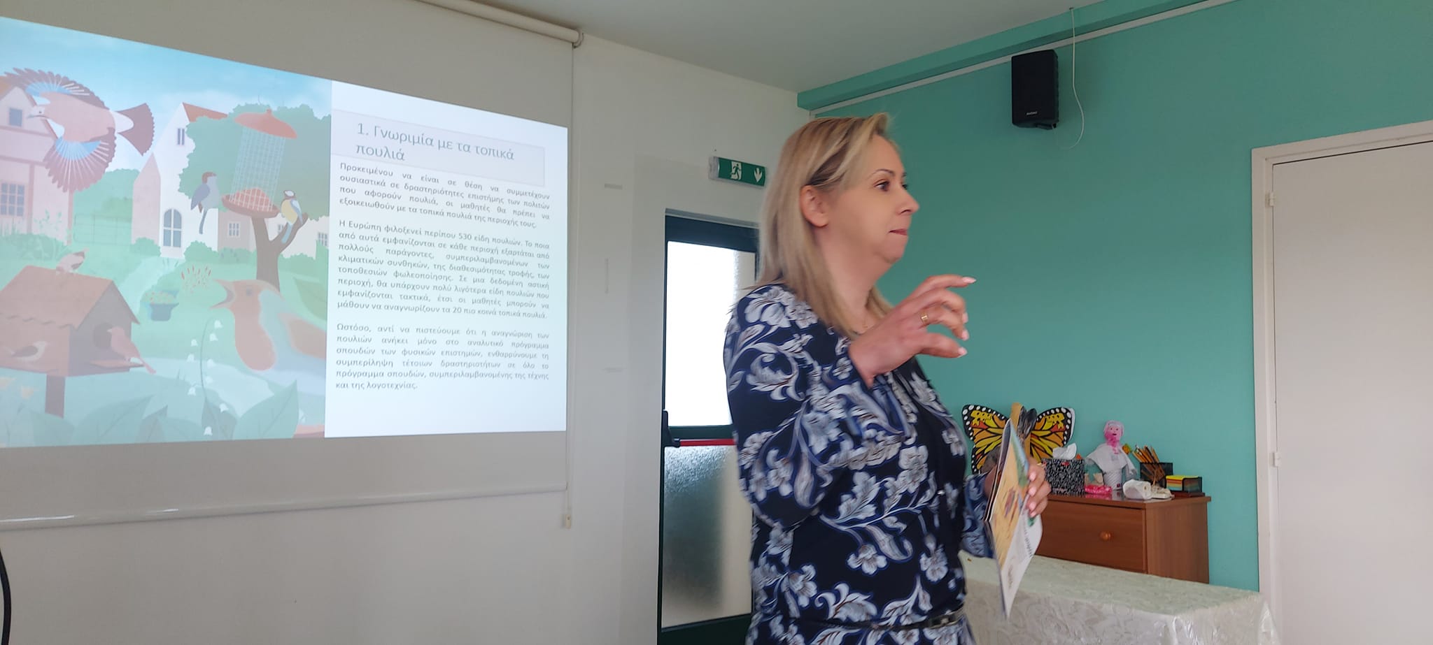 A lecturer stands in the lecture room. Behind her is a Powerpoint slide of a page from the EnviroCitizen project activity sheets for schools. The text on the screen is in Greek