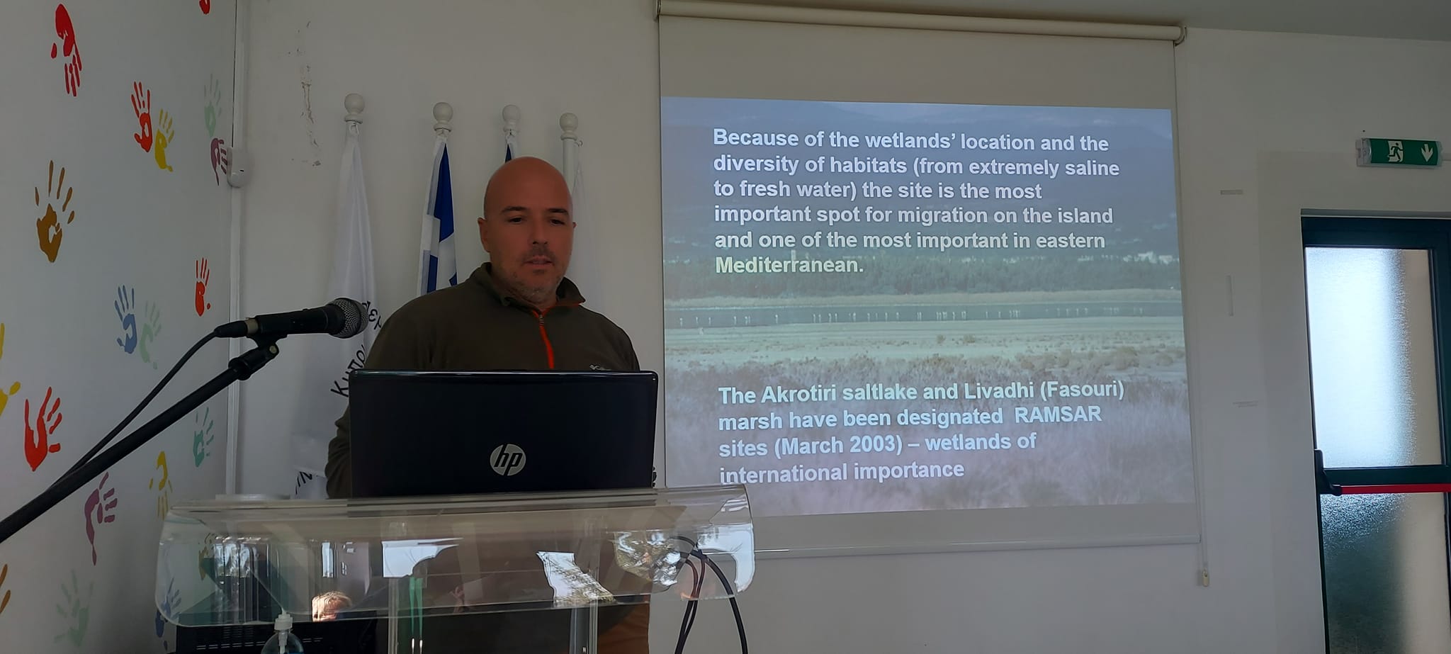 A man stands at a lectern with a Powerpoint slide in English on the screen behind him.